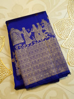 Load image into Gallery viewer, A folded royal blue silk saree with intricate gold zari woven motifs depicting figures and a detailed border, displayed on a beige patterned surface with decorative gold leaf accents.
