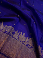 Load image into Gallery viewer, A close-up of a royal blue silk fabric, likely a saree, featuring intricate gold zari brocade work along the border with traditional figures and motifs.
