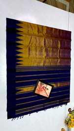Load image into Gallery viewer, A dark navy blue and gold striped silk saree is draped on a white wall, with a small framed picture placed on the fabric. The background shows a white wall with some minor home decor elements visible at the edges.
