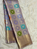 Load image into Gallery viewer, A folded luxury silk saree with a lavender and gold base, featuring woven square motifs in turquoise, green, and orange, displayed on a cream satin fabric background.
